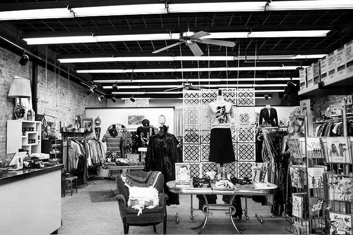 The inside of a retail clothing store, featuring exposed brick walls, a checkout counter, a sitting area and racks of clothes. Black and white photo.