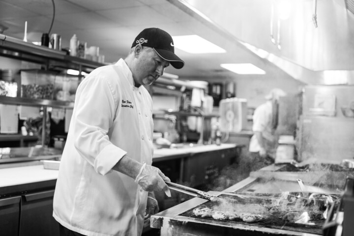 Chef grilling patties in a professional kitchen, wearing a cap and gloves. Black and white.