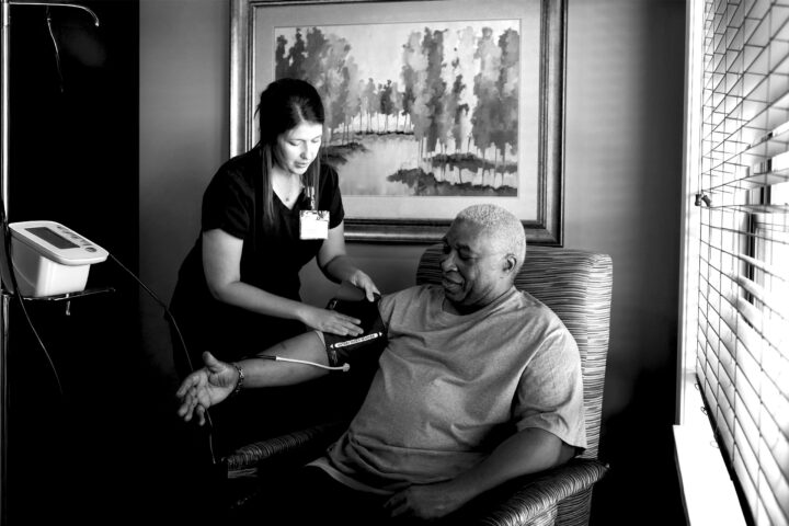 A caregiver checking the blood pressure of an older adult male sitting in an armchair near a window of a well-lit room. Black and white.