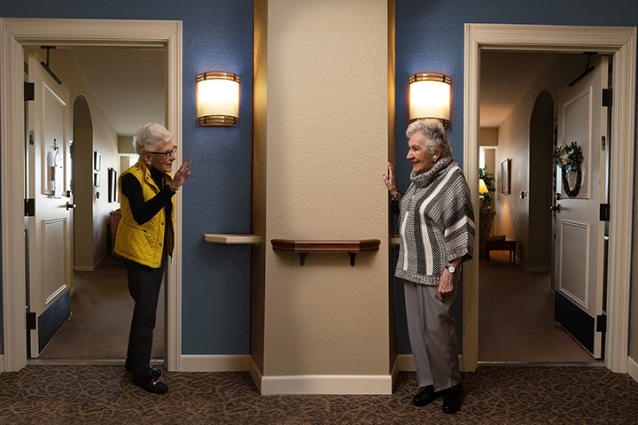 Two older women greet each other from separate neighboring doorways in a hallway, smiling and waving.