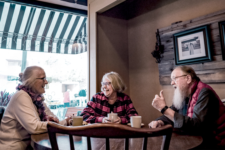 Three elderly people smiling and talking at a table in a cafe, with coffee cups in front of them.