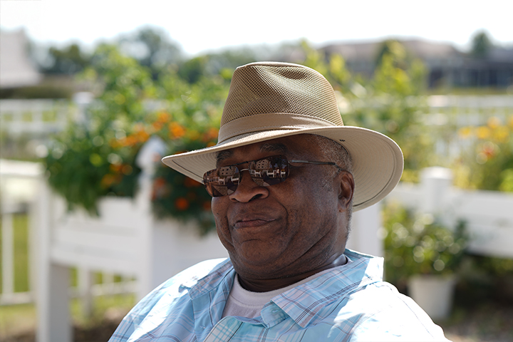 An older gentleman wearing a stetson hat and sunglasses smiles with raised garden beds behind him on a sunny day.