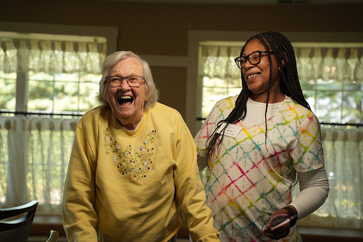 A caregiver supports a smiling elderly woman, both laughing and enjoying their time together.