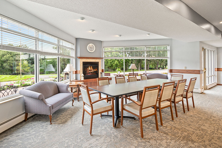 Spacious room with a long table, chairs, a couch, large windows, and a fireplace with a clock above.