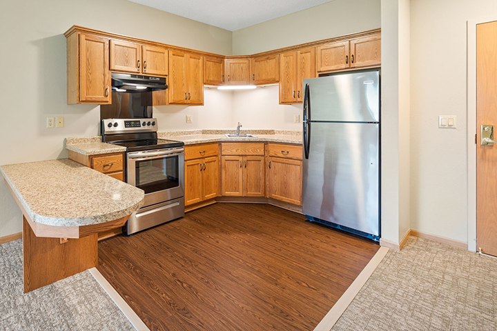 Modern kitchen in an Assisted Living Apartment with wood cabinets, stainless steel appliances, and a corner peninsula counter.