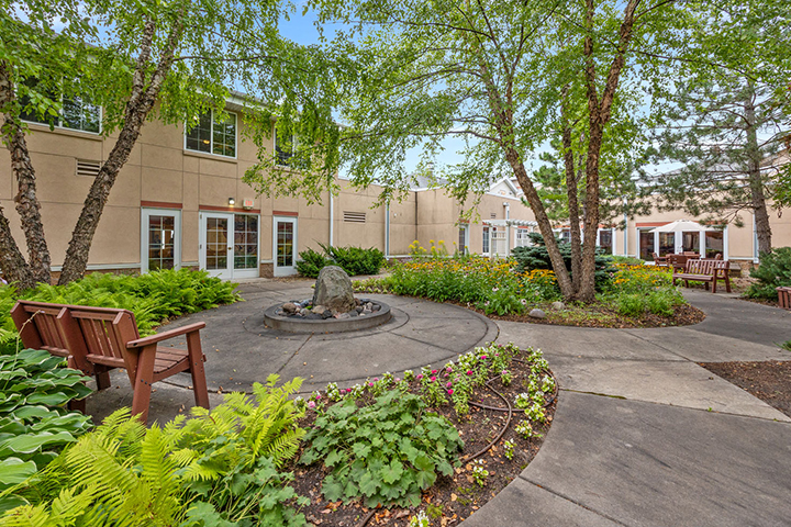 A landscaped courtyard with flowers, a fountain, benches, trees, and a beige building in the background.