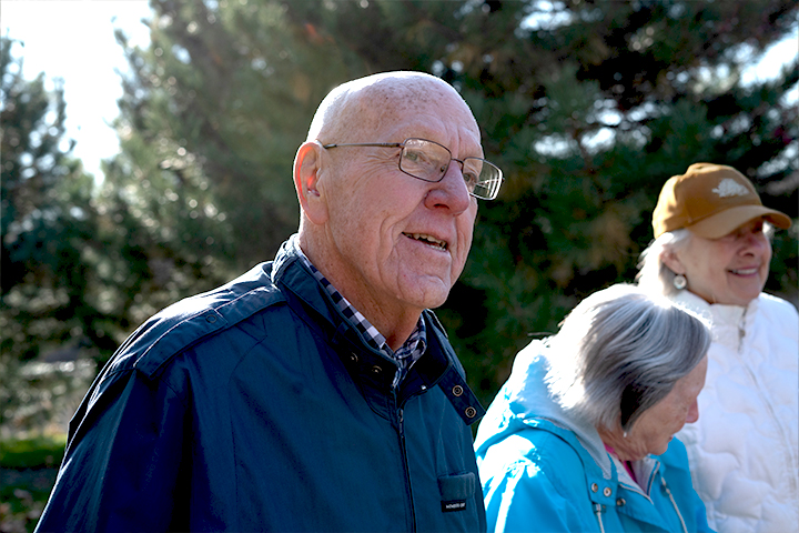 An bald man with eyeglasses and two elderly women wearing fall jackets walk side-by-side next to pine trees.