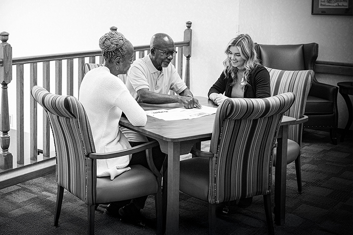 Three people sit at a table, smiling and discussing papers in a bright, comfortable room.