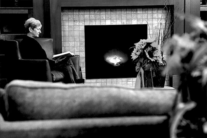 A woman sits in a leather chair reading a book next to a gas fireplace. Black and white.