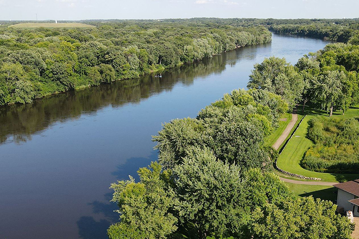 An aerial view of a river surrounded by lush, green trees and a paved path through parkland.
