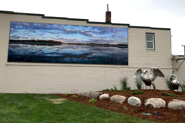 Swan sculptures stand in a garden near a painted flat-roof brick building adorned by a mural of a lake view at sunset.