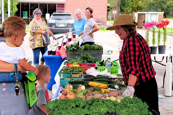 A woman and her two kids gather in front of a table covered in fresh produce as they talk to the woman farmer wearing a sun hat and plaid shirt.