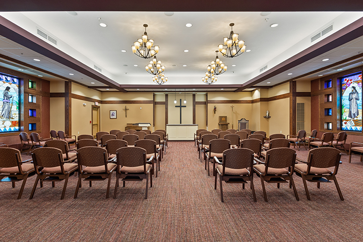 Chairs arranged in rows facing an altar with a cross in a well-lit chapel adorned with elegant chandeliers.