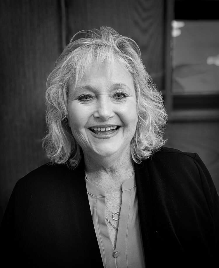 A woman with medium-length hair smiles for an indoor portrait wearing a blouse and dark jacket. Black and white.