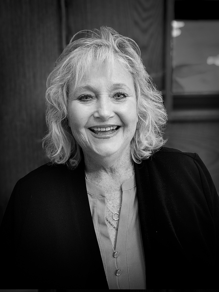 A woman with medium-length hair smiles for an indoor portrait wearing a blouse and dark jacket. Black and white.