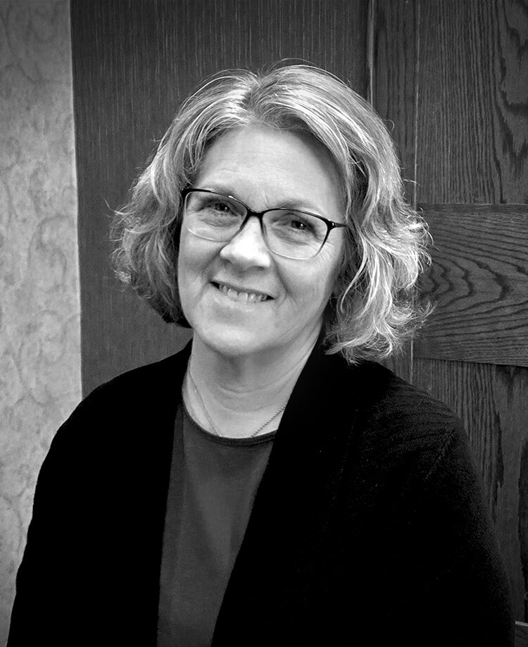 A woman with medium-length hair and eye glasses smiles for an indoor portrait wearing a blouse and dark jacket. Black and white.