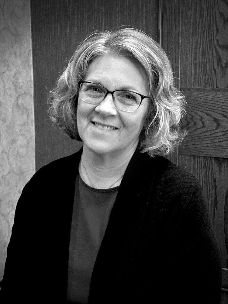 A woman with medium-length hair and eye glasses smiles for an indoor portrait wearing a blouse and dark jacket. Black and white.