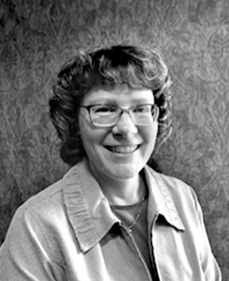 A woman with short-cropped hair and eyeglasses smiles for an indoor portrait wearing a blouse and light jacket. Black and white.