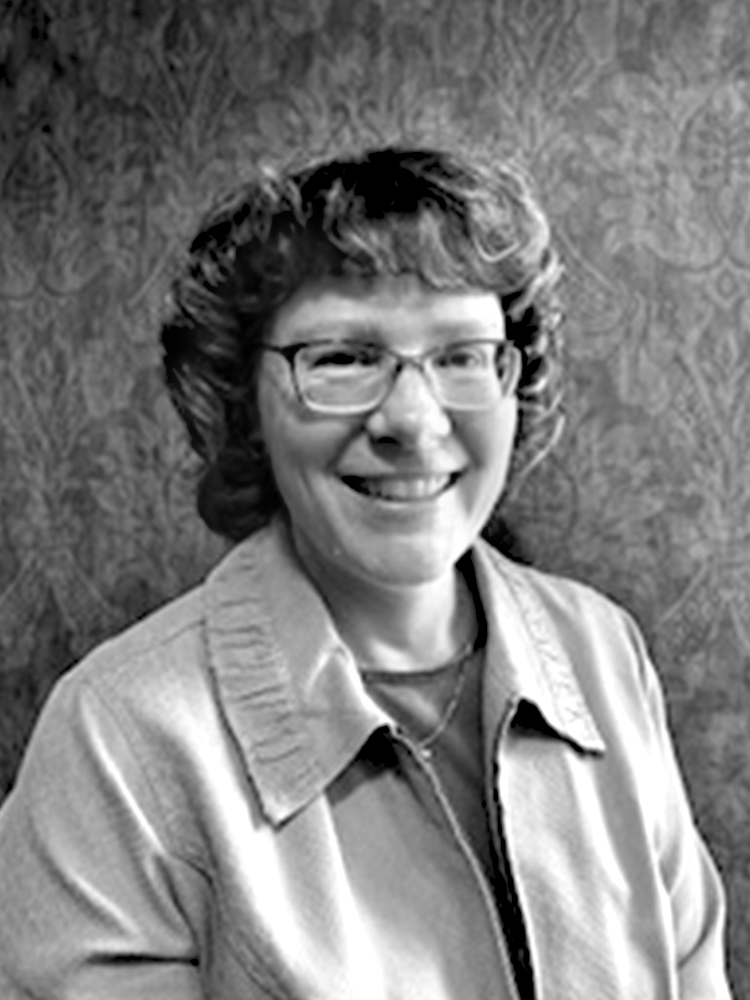 A woman with short-cropped hair and eyeglasses smiles for an indoor portrait wearing a blouse and light jacket. Black and white.