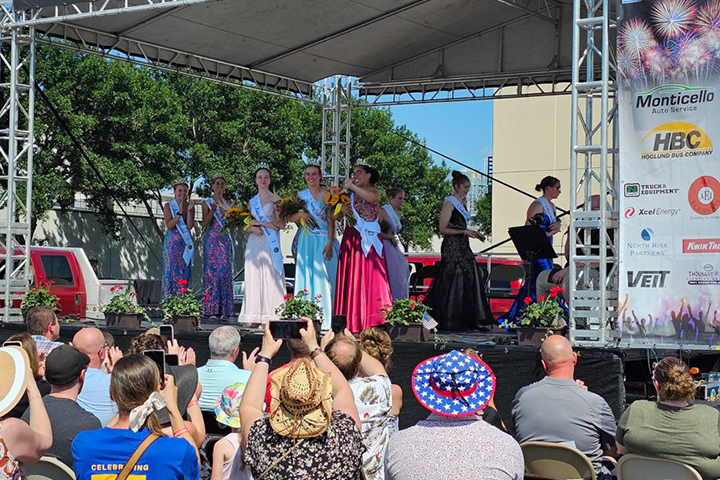 Winners of female pageantry, wearing gowns and crowns, wave to an audience from a stage during the Monticello Riverfest.