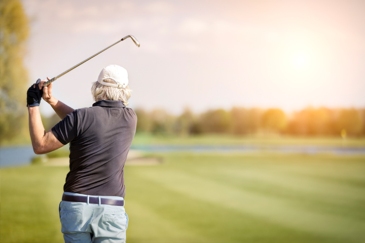 A man with medium-length silver hair, blue pants and black polo shirt, holds his follow-through golf swing while watching his ball down the fairway on a sunny day.