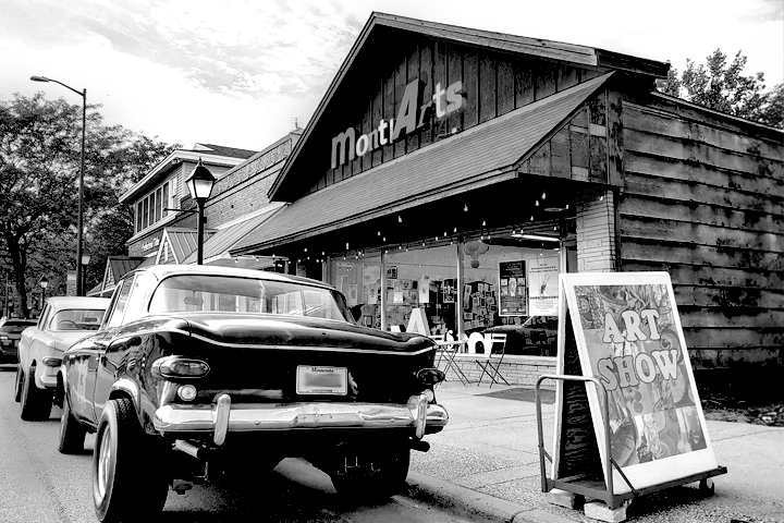 Custom art cars from the '60s and '70s era parked outside a gallery shop called MontiArts. A sandwich board on the sidewalk features the words Art Show. Black and white.