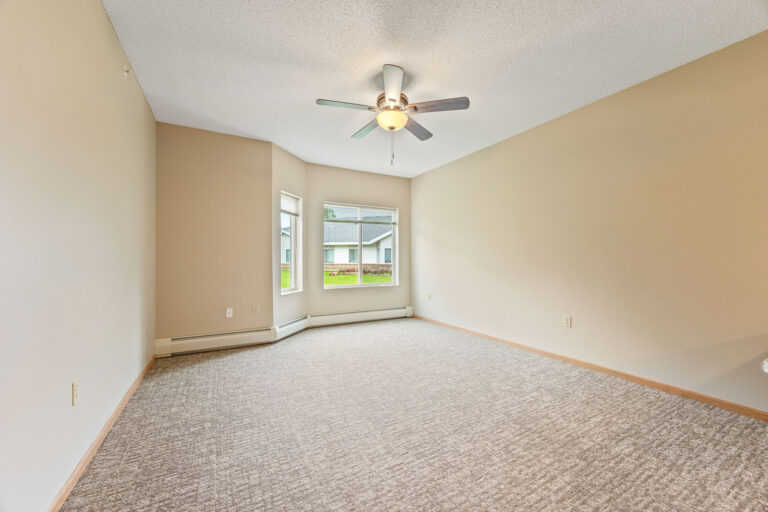 Empty beige apartment with carpeted floor, ceiling fan, large windows, and views of lawn and neighboring houses.