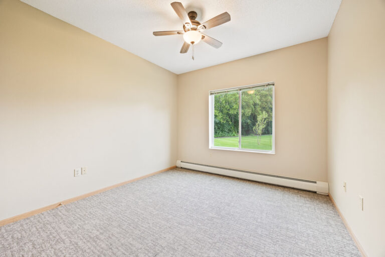 Empty beige room with a ceiling fan, large window, and carpeted floor, overlooking green trees outside.