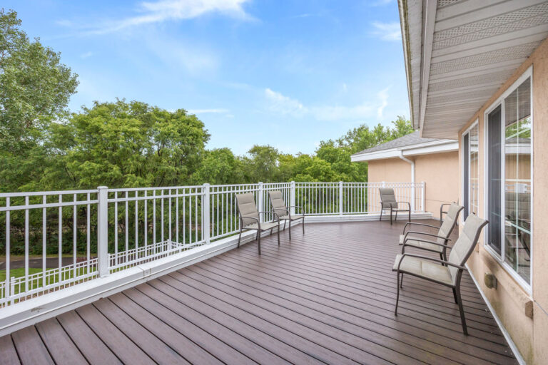 Spacious assisted living deck with chairs, white railing, and a view of green trees under a blue sky.