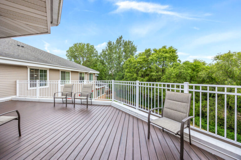 Spacious assisted living deck with chairs, white railing, and a view of green trees under a blue sky.