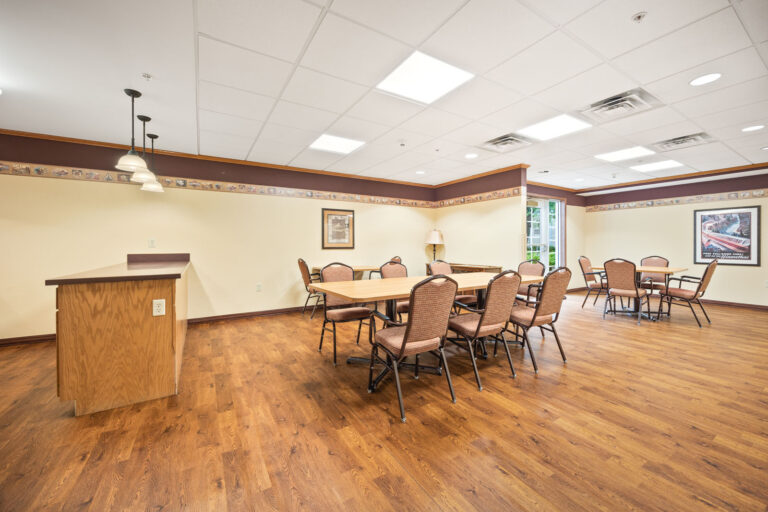 A spacious community room with tables, chairs, wood flooring, and bright overhead lighting.