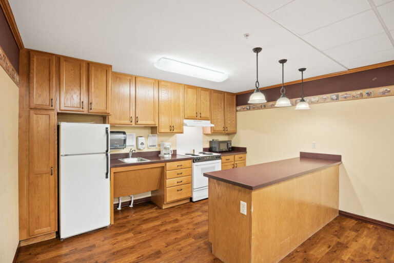 Assited living kitchen with wood cabinets, white appliances, island, and hanging lights on a wood floor.