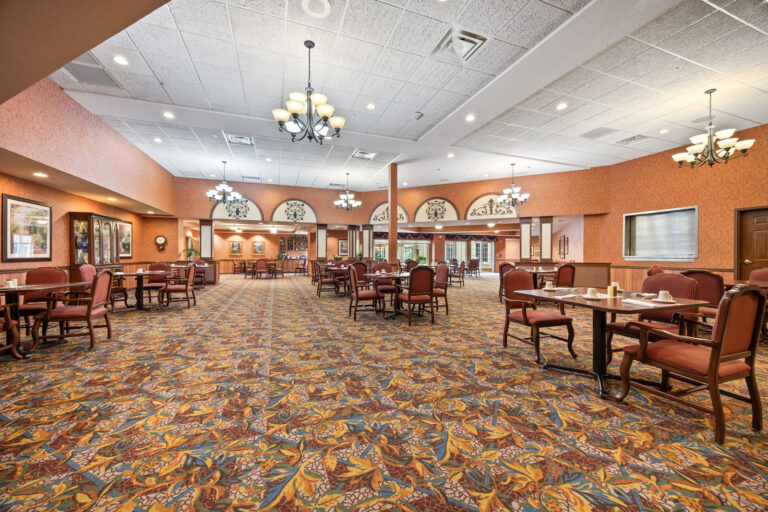 Spacious dining room with patterned carpet, chandeliers, and tables set with red chairs and place settings.