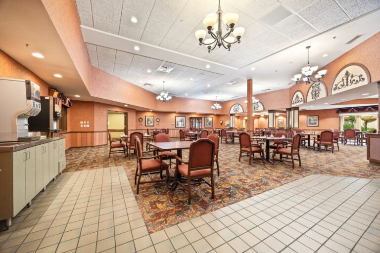 Assisted Living dining room with patterned carpet, chandeliers, and tables set with red chairs and place settings.