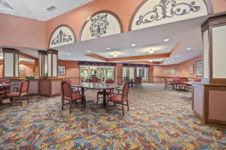A spacious dining room with round tables, red chairs, ornate carpet, and decorative wall accents.