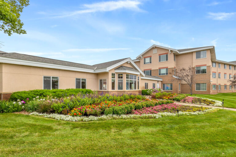Court assisted living building entrance with large windows, surrounded by a colorful flower garden and green lawn under a blue sky.