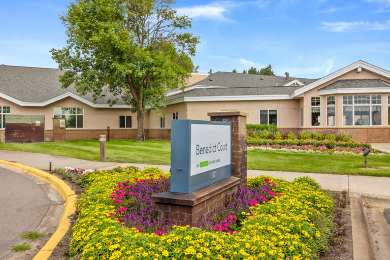 Exterior of Benedict Court building with a sign surrounded by yellow and purple flowers and green trees.