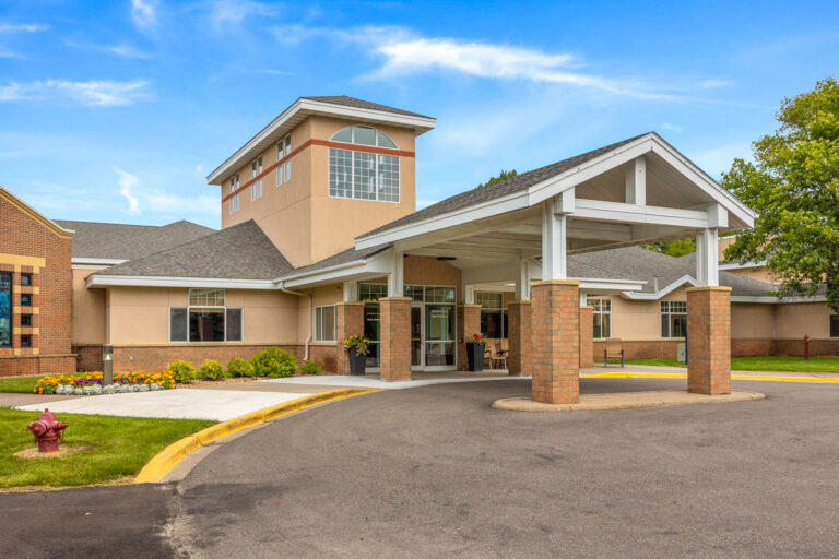 Court assisted living single-story building with a covered entrance, surrounded by greenery and a clear blue sky.