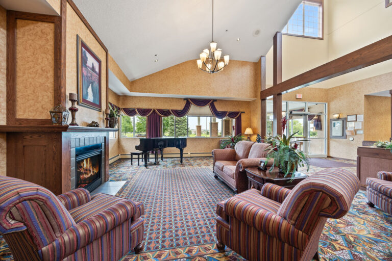 A cozy lounge with striped chairs, a fireplace, a grand piano, and large windows letting in natural light.