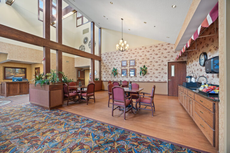 Lobby area with tables, chairs, food counter, floral carpet, and high ceilings with large windows.