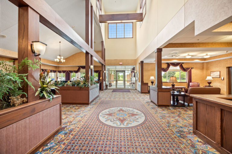 Spacious lobby with patterned carpet, large windows, plants, and wooden accents, with seating areas on the side.
