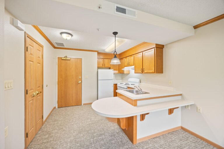 Assisted living kitchen with wood cabinets, white appliances, and a curved peninsula counter in a carpeted area.