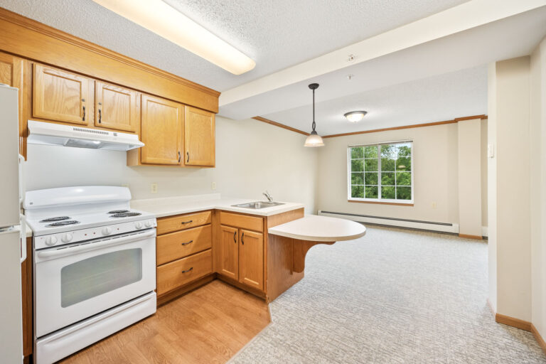 Assisted living kitchen with wood cabinets, white appliances, and a counter facing a bright living area with a large window.