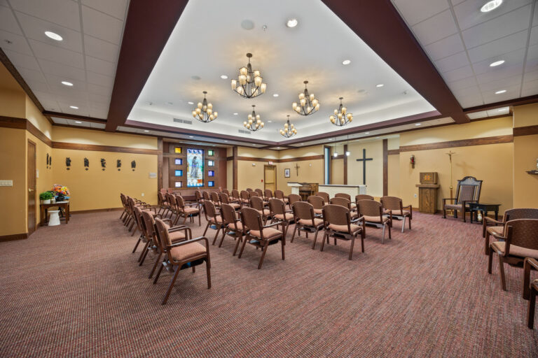 Chairs arranged in rows facing an altar with a cross in a well-lit chapel adorned with elegant chandeliers.