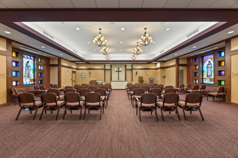 Chairs arranged in rows facing an altar with a cross in a well-lit chapel adorned with elegant chandeliers.