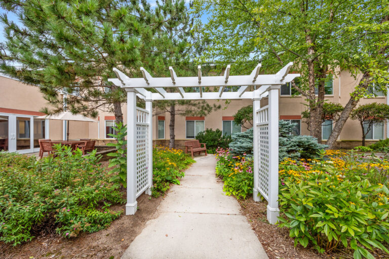 White garden pergola over sidewalk path, surrounded by green plants and flowers near a building with windows.