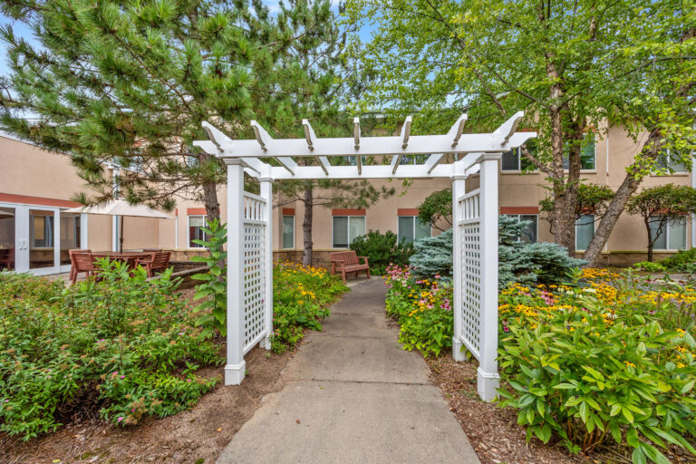 White garden arbor over a path, surrounded by green plants and flowers near a building with windows.