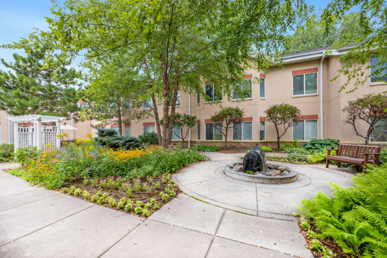 A landscaped courtyard with flowers, a fountain, benches, trees, and a beige building in the background.