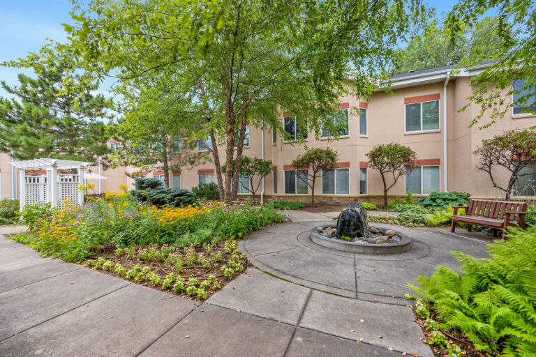 A landscaped courtyard with flowers, a fountain, benches, trees, and a beige building in the background.