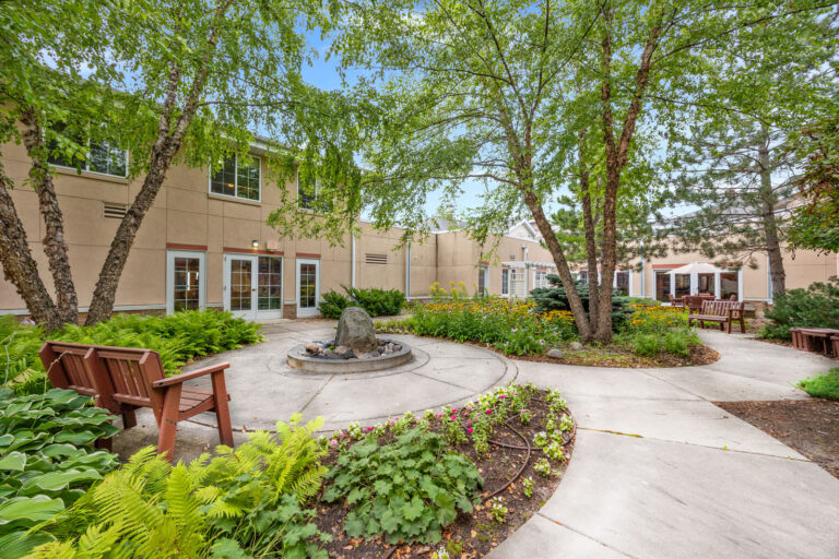 A landscaped courtyard with flowers, a fountain, benches, trees, and a beige building in the background.